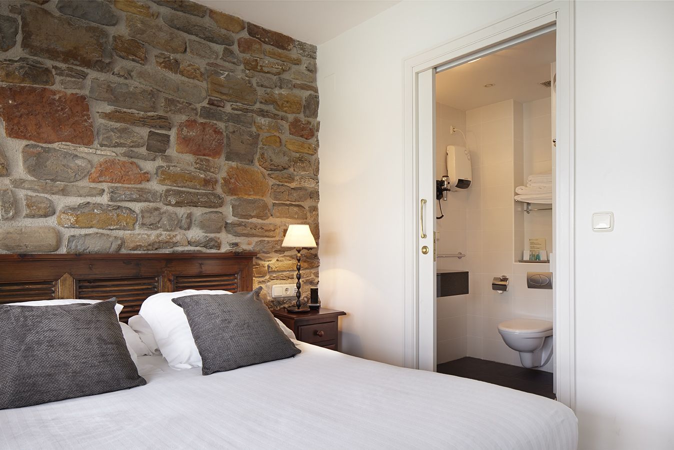 A bedroom with a stone wall, a bed with pillows, a bedside table with a lamp, and an open door to a bathroom.