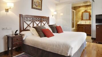 A bedroom with a wooden headboard, a bedside table, a nightstand, a bed with white bedding and red pillows, a wall-mounted lamp, and a painting.