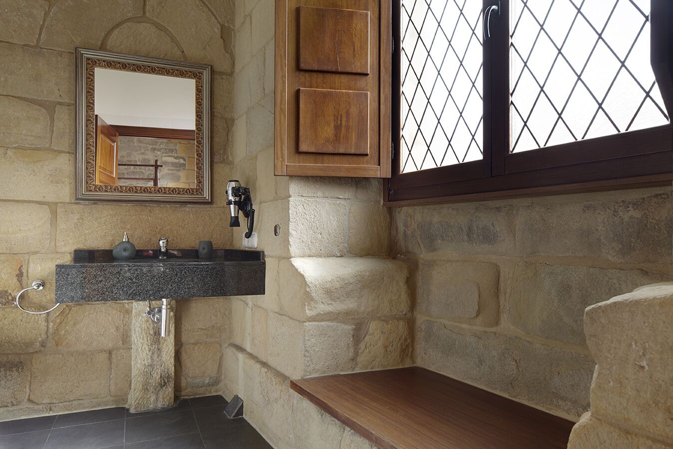 A stone-walled bathroom with a granite sink, a mirror, and a wooden shelf.
