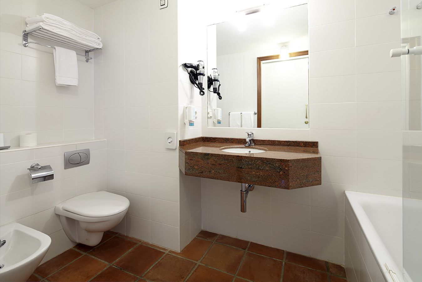 Une salle de bain avec des toilettes blanches, un bidet, un lavabo doté d’un plan de travail en granit et un porte-serviettes avec des serviettes pliées.