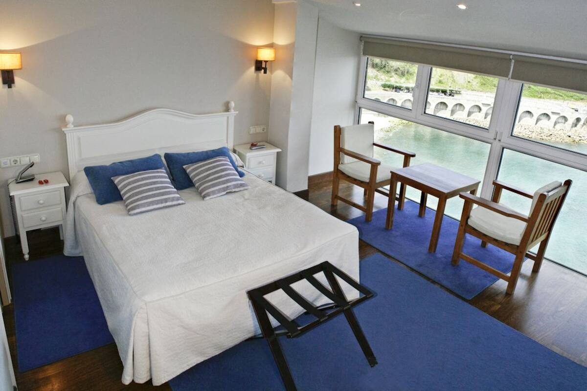 Une chambre à coucher comprenant un lit, des tables de chevet, une chaise, une petite table et une vue sur un pont enjambant une étendue d’eau.