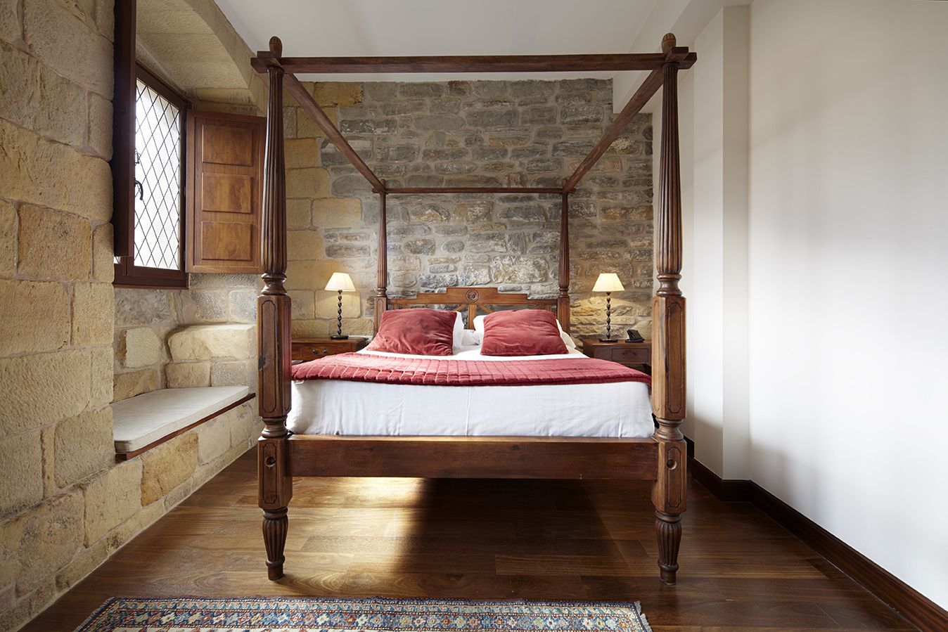 A four-poster bed with a stone wall backdrop, wooden flooring, and a small bench by the window.
