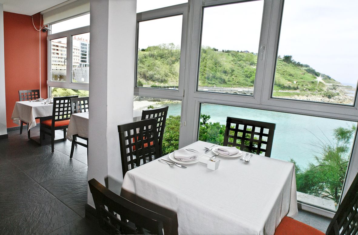 A dining area with tables set for two, large windows with a view of a green hillside and a body of water.