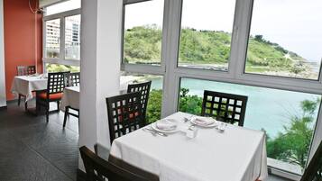 A dining area with tables set for two, large windows with a view of a green hillside and a body of water.