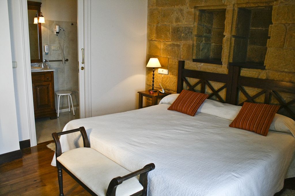 Un dormitorio con una cama grande, cabecera de madera, dos almohadas, una mesita de noche con lámpara, una silla de madera y un baño visible a través de una puerta abierta.