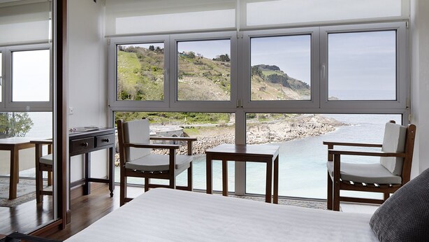 A bedroom with a large window overlooking a rocky beach and hills. - Saiaz Getaria Hotela (Getaria)