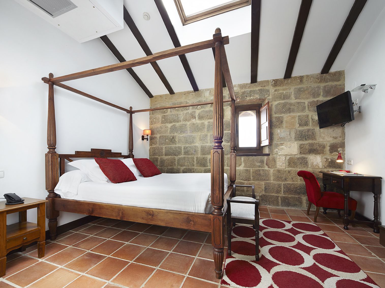 A four-poster bed with a stone wall backdrop, a desk with a chair, and a TV mounted on the wall.