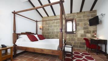 A four-poster bed with a stone wall backdrop, a desk with a chair, and a TV mounted on the wall.