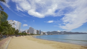 A beach with a sandy shore, palm trees, and a cityscape with high-rise buildings in the background. - Paralia Hotel Nha Trang (Bac Nha Trang)