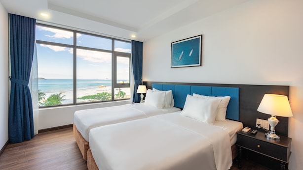 A hotel room with two beds, a large window with a view of the beach, a blue headboard, and a nightstand with a lamp. - Paralia Hotel Nha Trang (Bac Nha Trang)