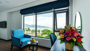 A room with a large window, a blue armchair, a small table with a fruit bowl, and a vase of flowers. - Paralia Hotel Nha Trang (Bac Nha Trang)