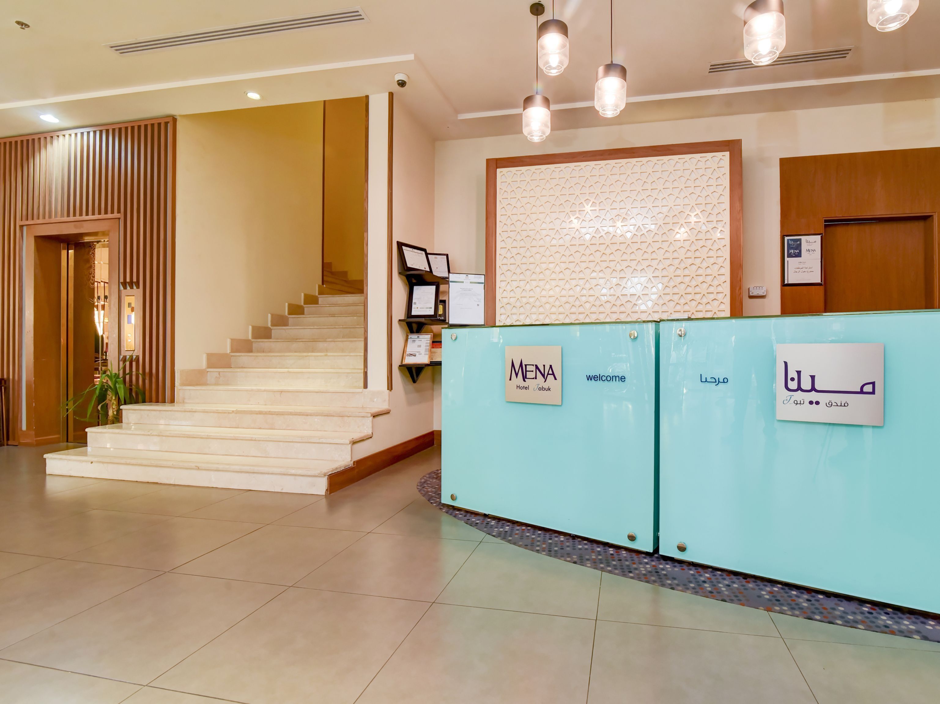 Zona de recepción de un hotel con una escalera, mostrador de recepción y una pared con certificados enmarcados.
