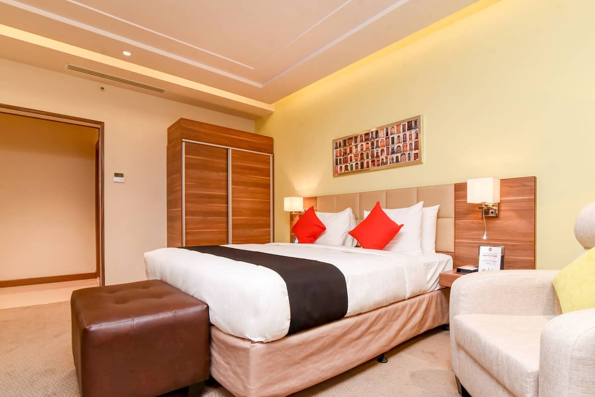 A hotel room with a large bed, a brown leather ottoman, a beige armchair, and a wooden wardrobe.
