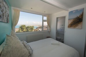 A bedroom with a large window offering a view of the sea and greenery. - Coast Accommodation (St Ives)