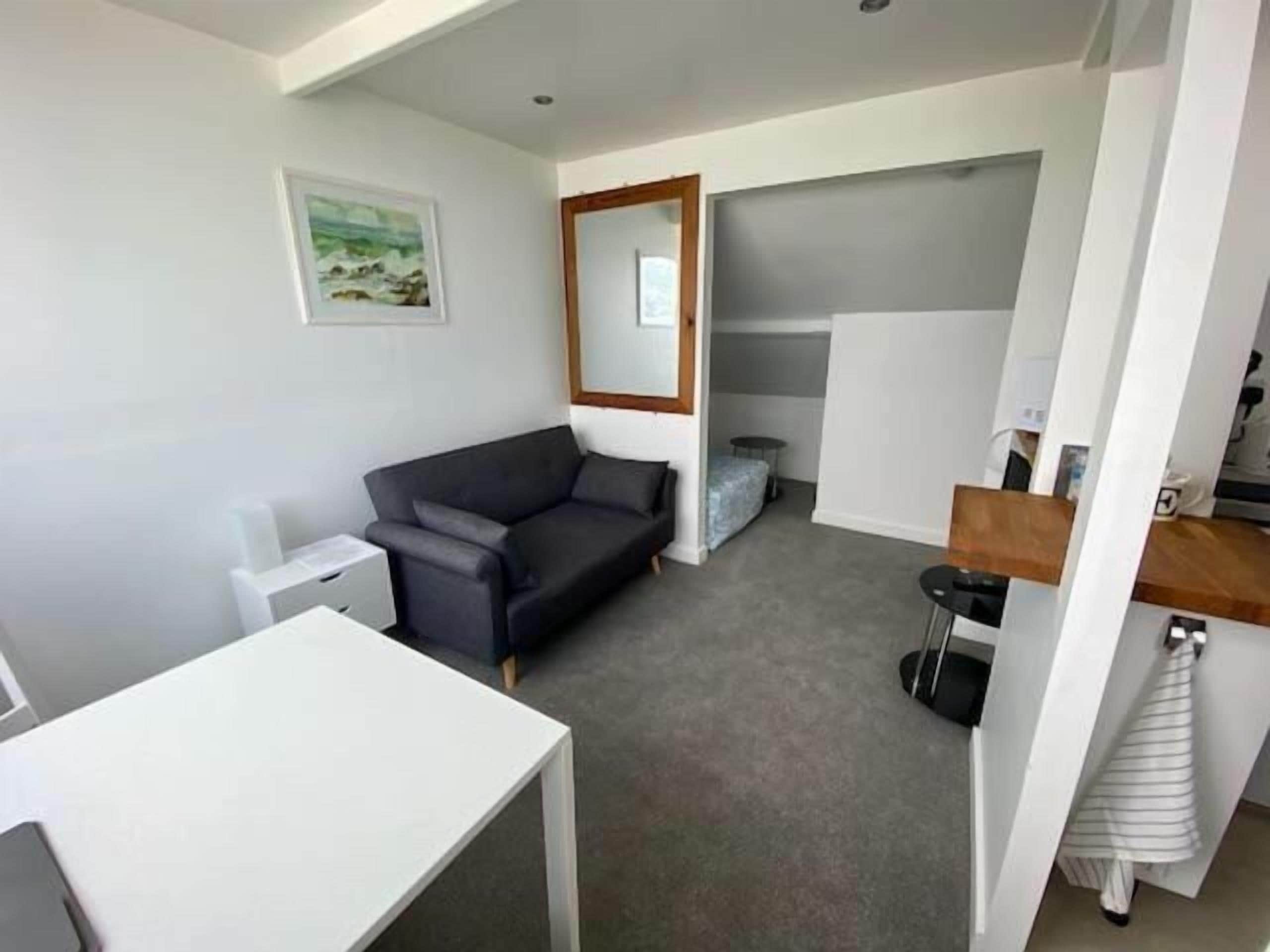 A compact living space with a sofa, a small table, a mirror, and a kitchenette area.
