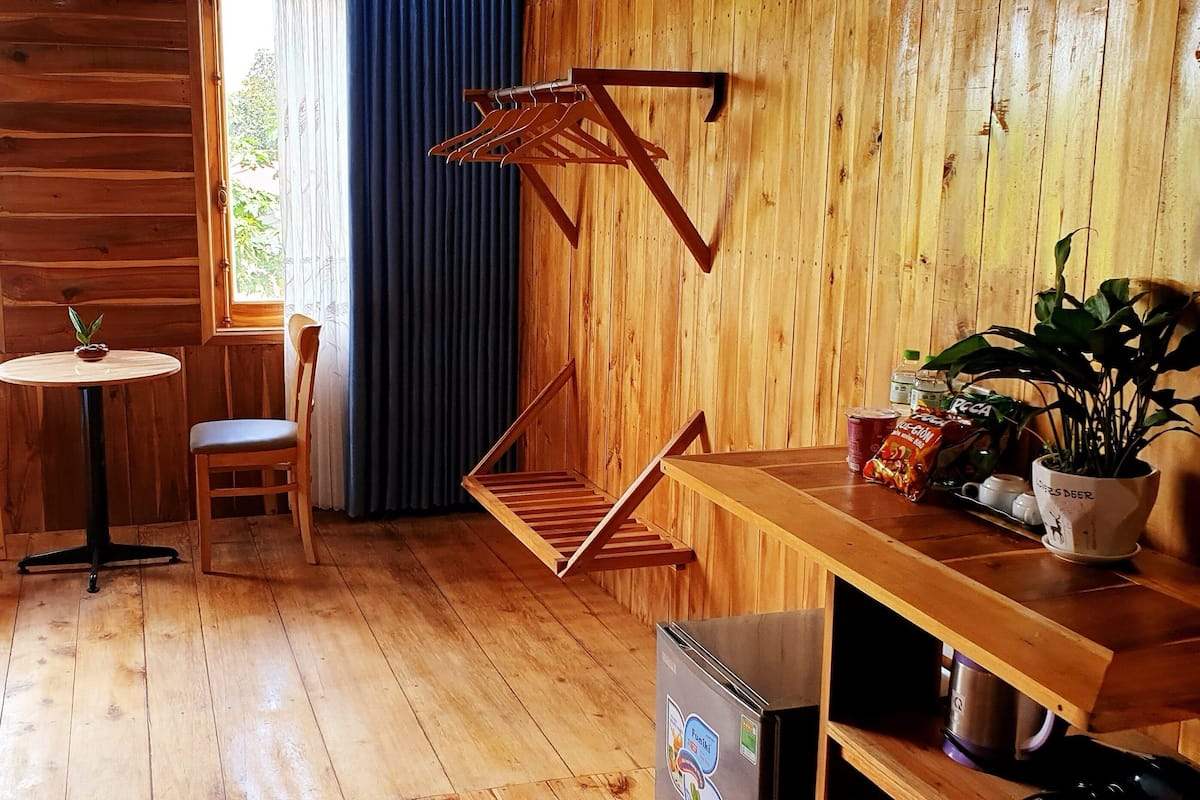 A wooden room with a bed, a table, a chair, a small table with a plant, and a hanging clothes rack.