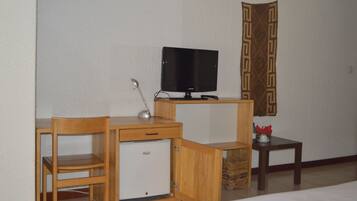 Una habitación funcional con un escritorio de madera, un refrigerador pequeño, un televisor y una mesita con un adorno decorativo.