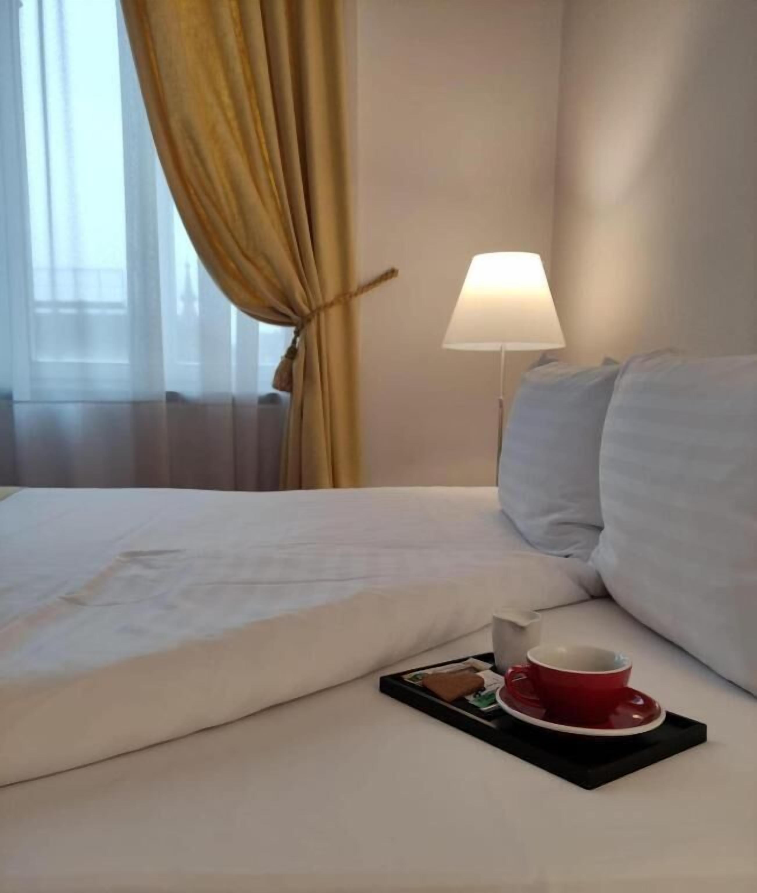 A hotel room with a neatly made bed, a bedside lamp, and a tray with a red cup and saucer.