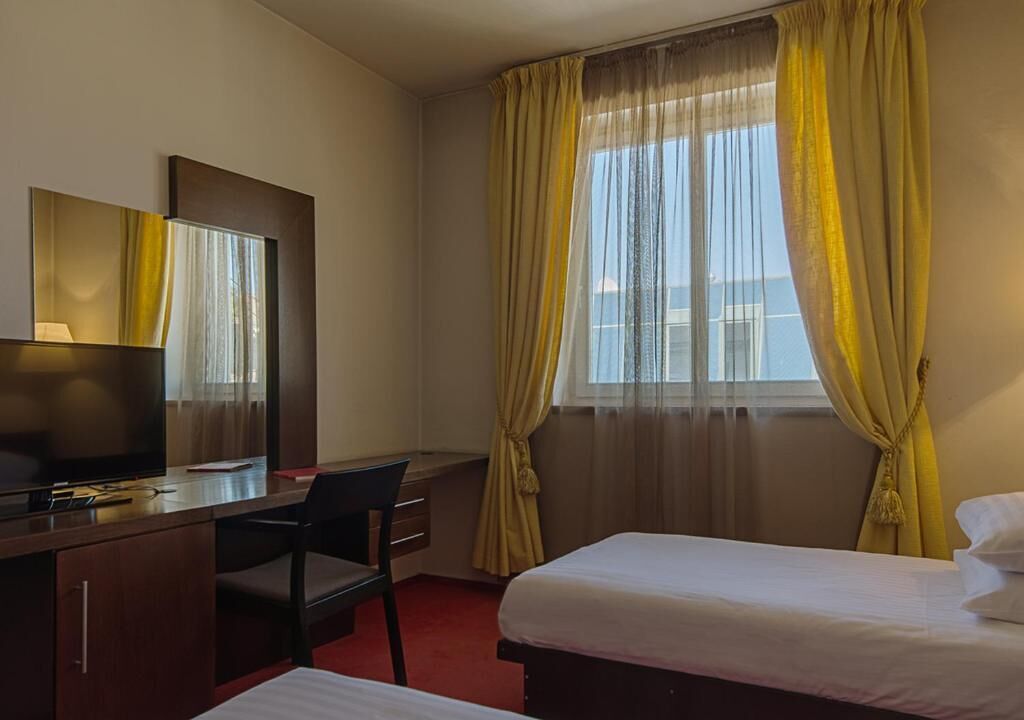A hotel room with a bed, a desk with a chair, a television, and a window with yellow curtains.