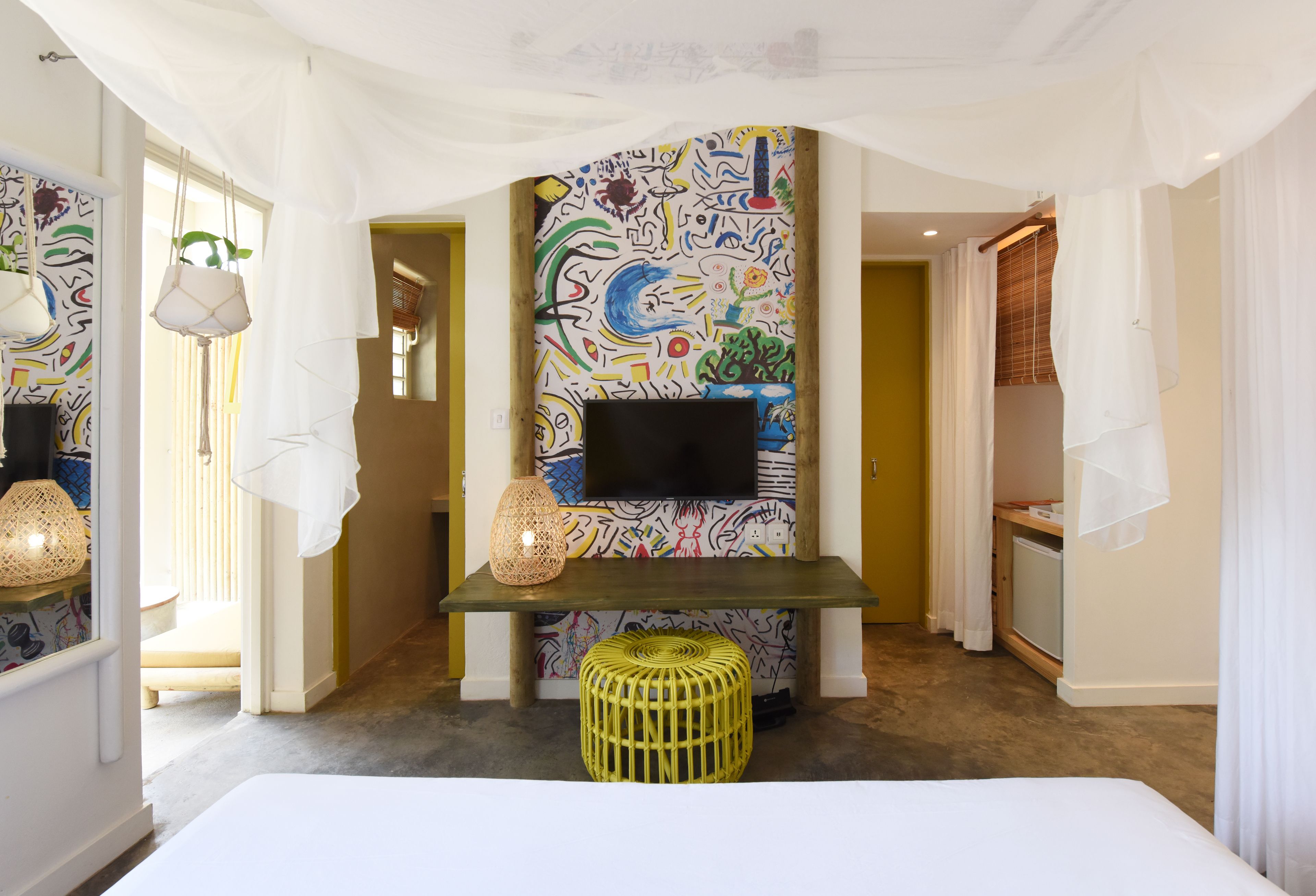 A hotel room with a colorful abstract mural, a flat-screen TV, a woven stool, and a bed.