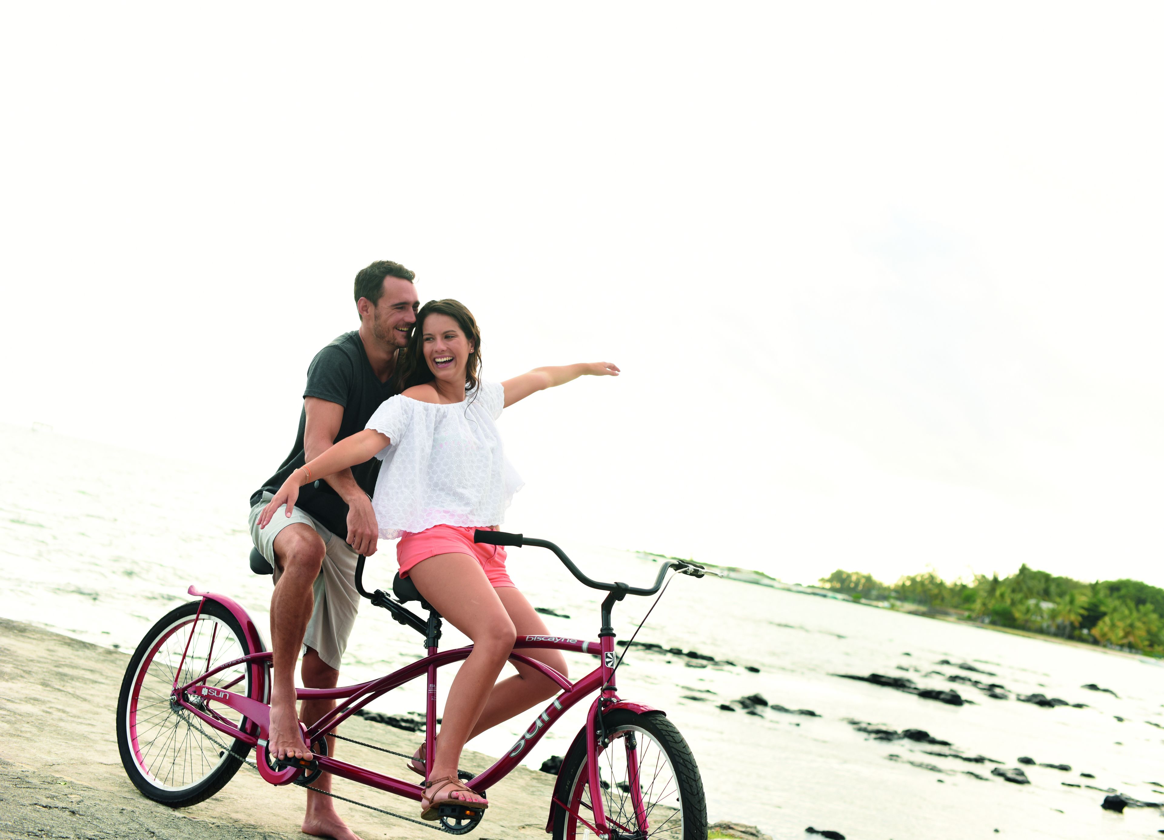 Un couple est assis sur un vélo rose, sur une plage, sous un ciel dégagé.