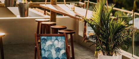A wooden pergola with ropes, wooden chairs, and tables on a rooftop terrace with potted plants.