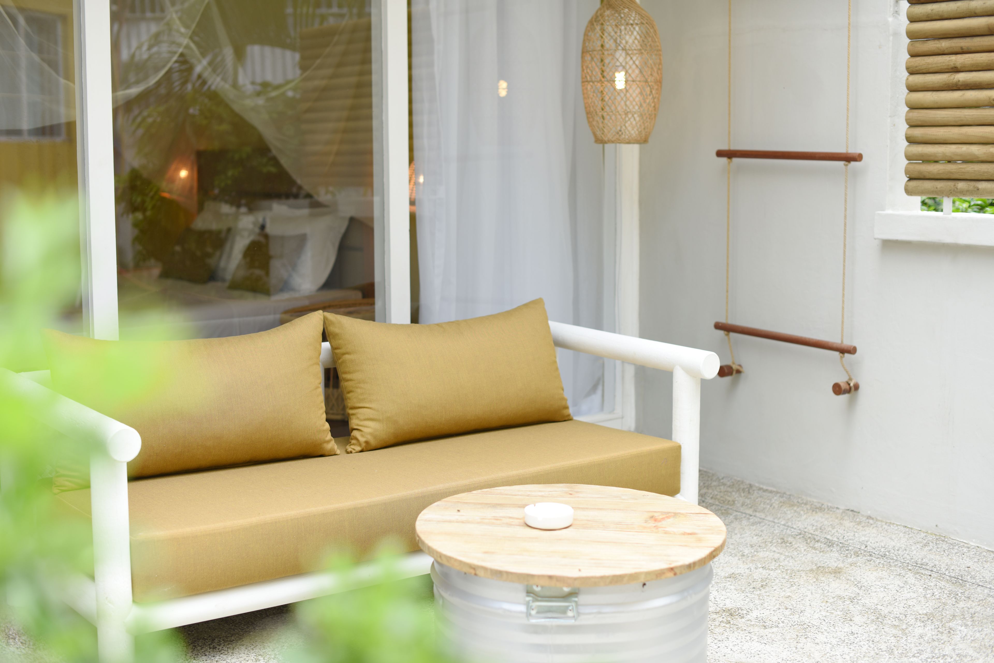 Un sofá blanco con un cojín beige, una mesa redonda de madera con un objeto blanco encima, y una lámpara colgante de mimbre.