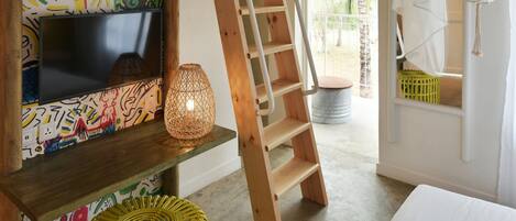 Una habitación con escalera de madera, un mural de pared colorido, una lámpara de pie tejida y una cama con ropa blanca.