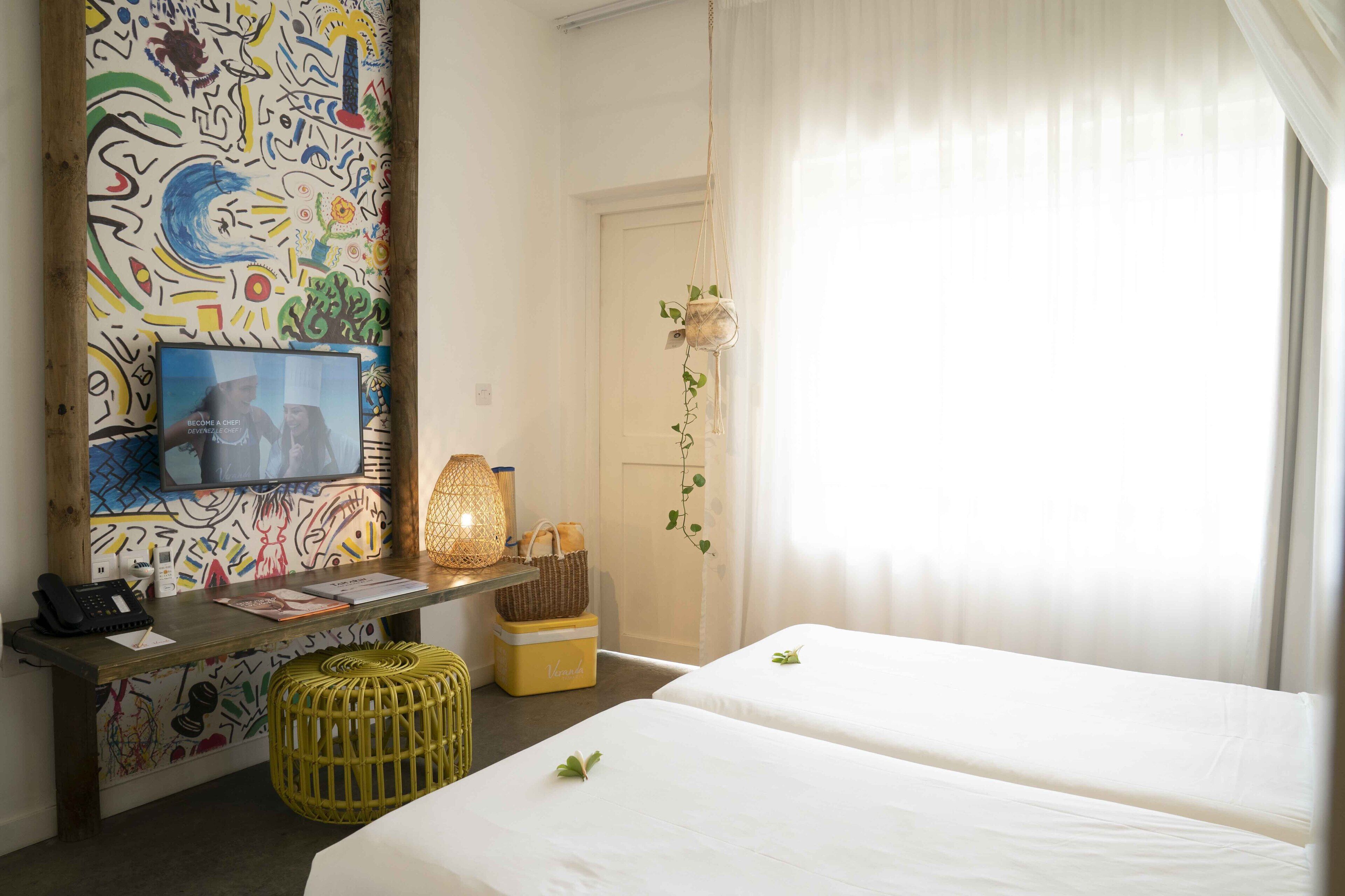 A hotel room with two beds, a desk with a phone and papers, a colorful abstract wall, and a window with sheer curtains.