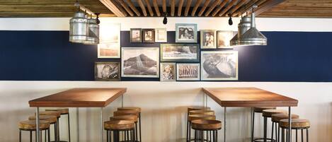 Zona de bar con mesas y taburetes de madera, cuadros en la pared y lámparas colgantes de metal.