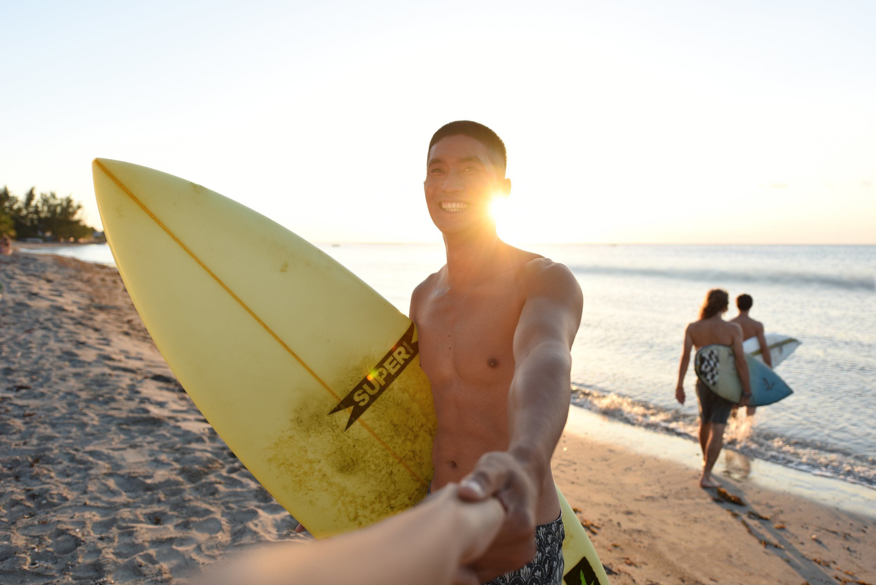 Cena de praia com uma pessoa segurando uma prancha de surf amarela e outra pessoa caminhando com uma prancha.