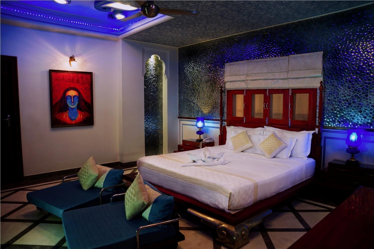 Una habitación de hotel moderna con una cama grande, un área de estar con sillas azules, una obra de arte colgada en la pared e iluminación ambiental.