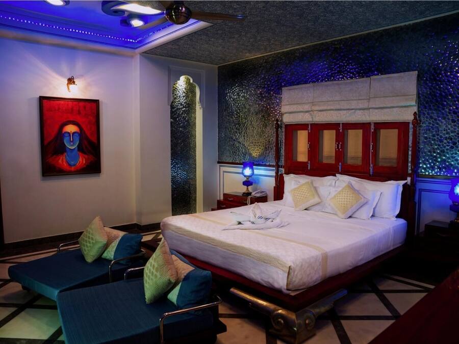 Una habitación de hotel moderna con una cama grande, un área de estar con sillas azules, una obra de arte colgada en la pared e iluminación ambiental.