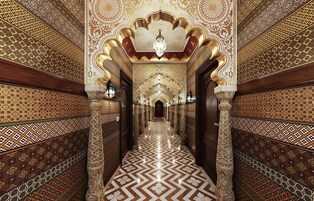 Um corredor com padrões geométricos elaborados nas paredes e no chão, colunas ricamente decoradas e um lustre.