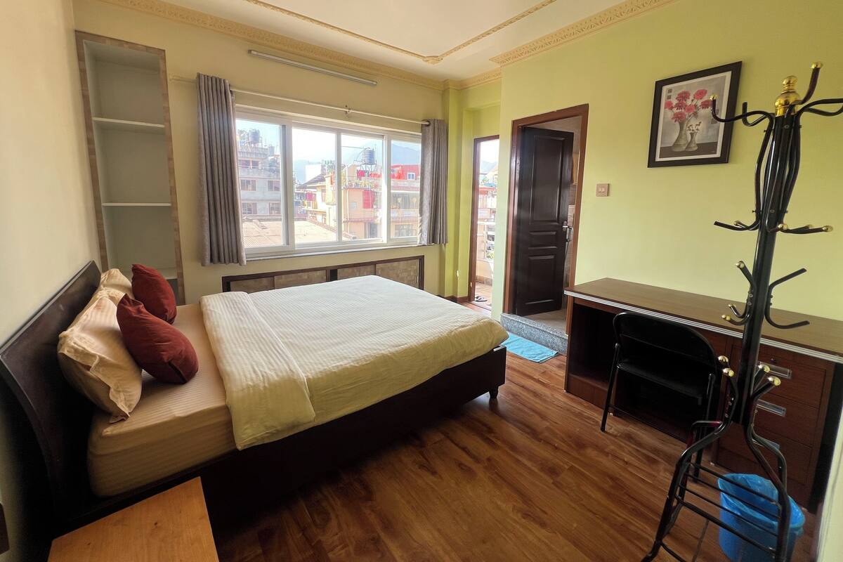 A bedroom with a bed, a desk, a chair, a coat rack, and a window with a view of buildings.