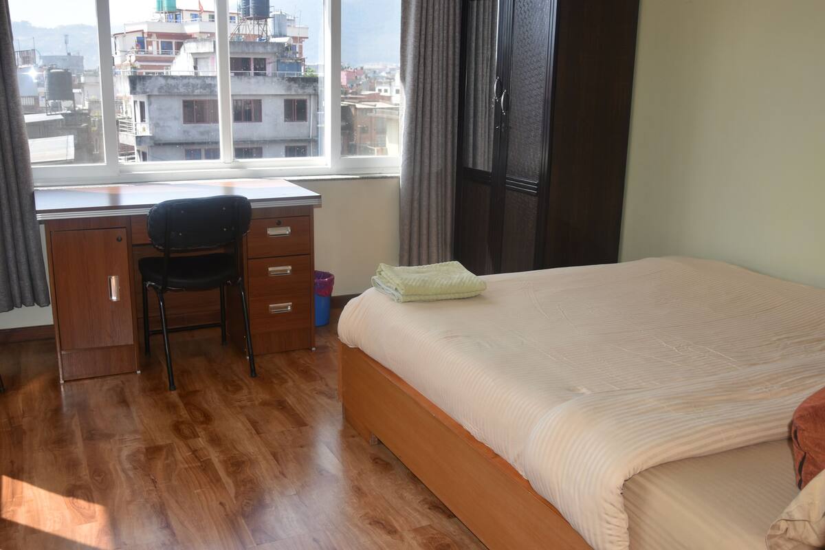A hotel room with a wooden desk, a chair, a bed with a beige bedspread, and a view of buildings through a large window.