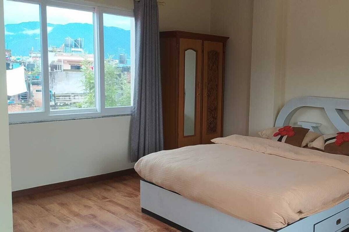 A bedroom with a bed, a nightstand, a wardrobe, and a window with a view of buildings and mountains.