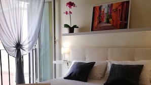 A hotel room with a bed, a nightstand, a lamp, a window with sheer curtains, a potted plant, and a painting on the wall. - Vetrina Suites (Rome)