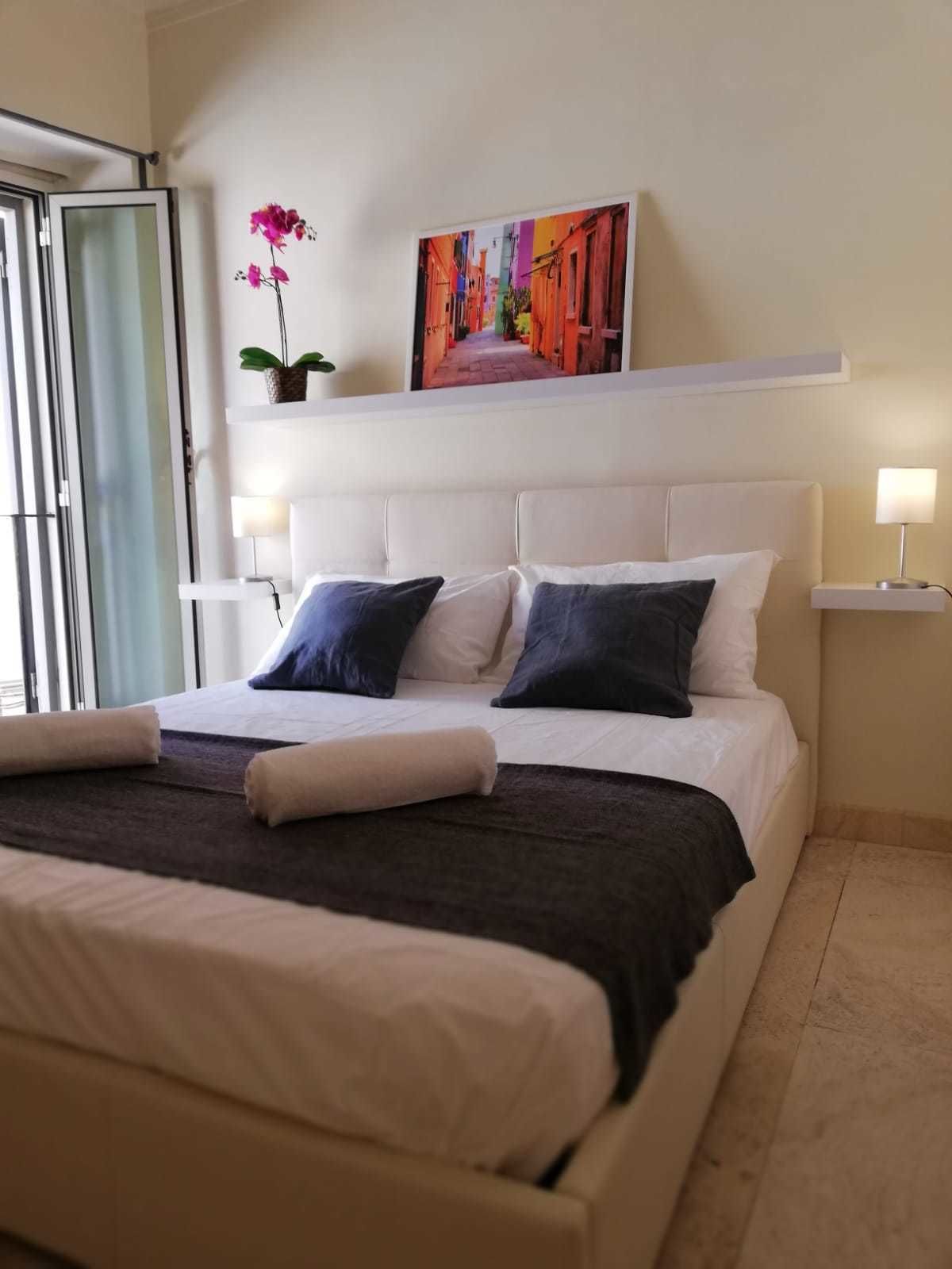 A neatly made bed with pillows, a bedside table with a lamp, a painting above the bed, and a potted plant on a shelf.