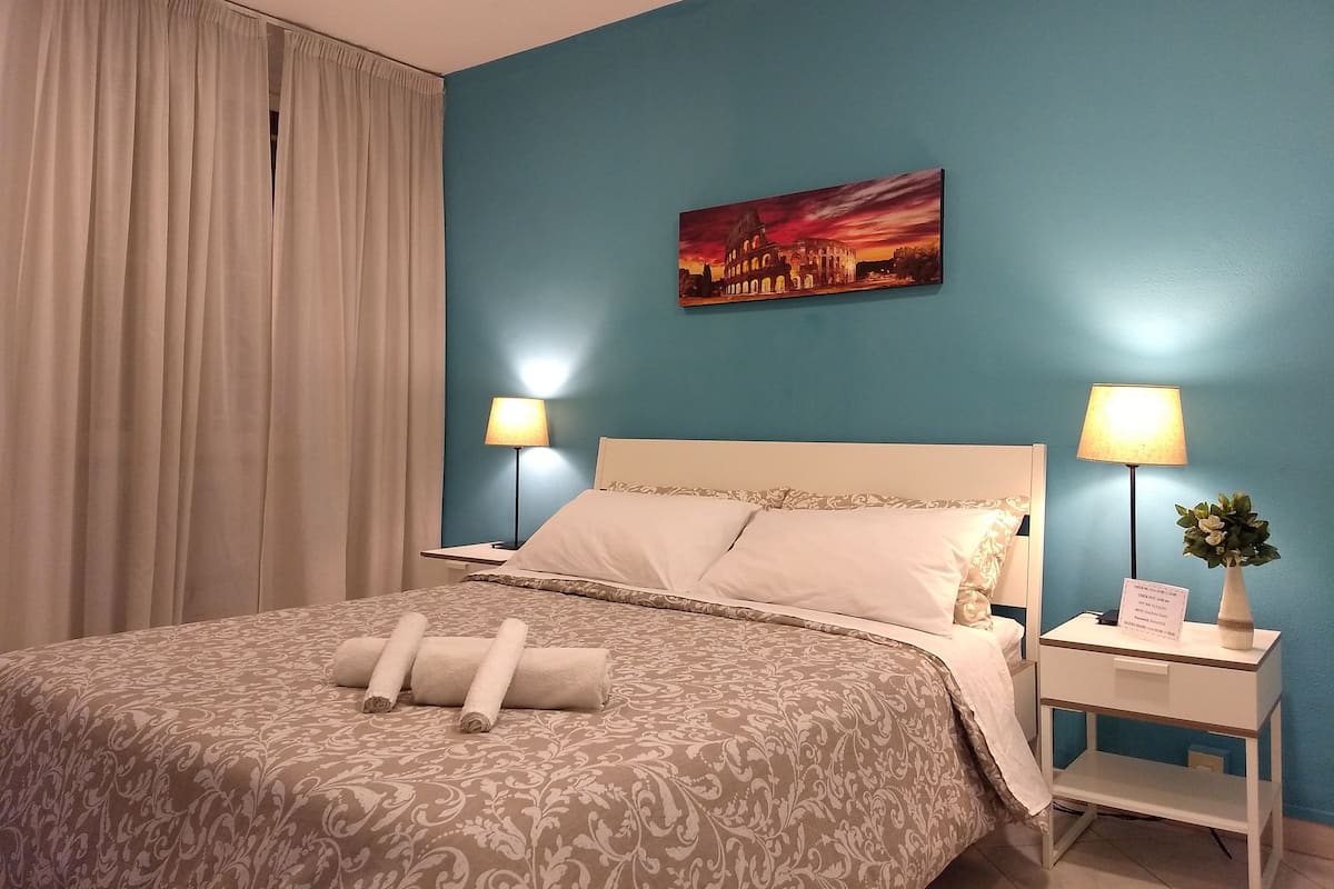 A hotel room with a bed, two bedside tables with lamps, a painting of a cityscape, and a window with curtains.
