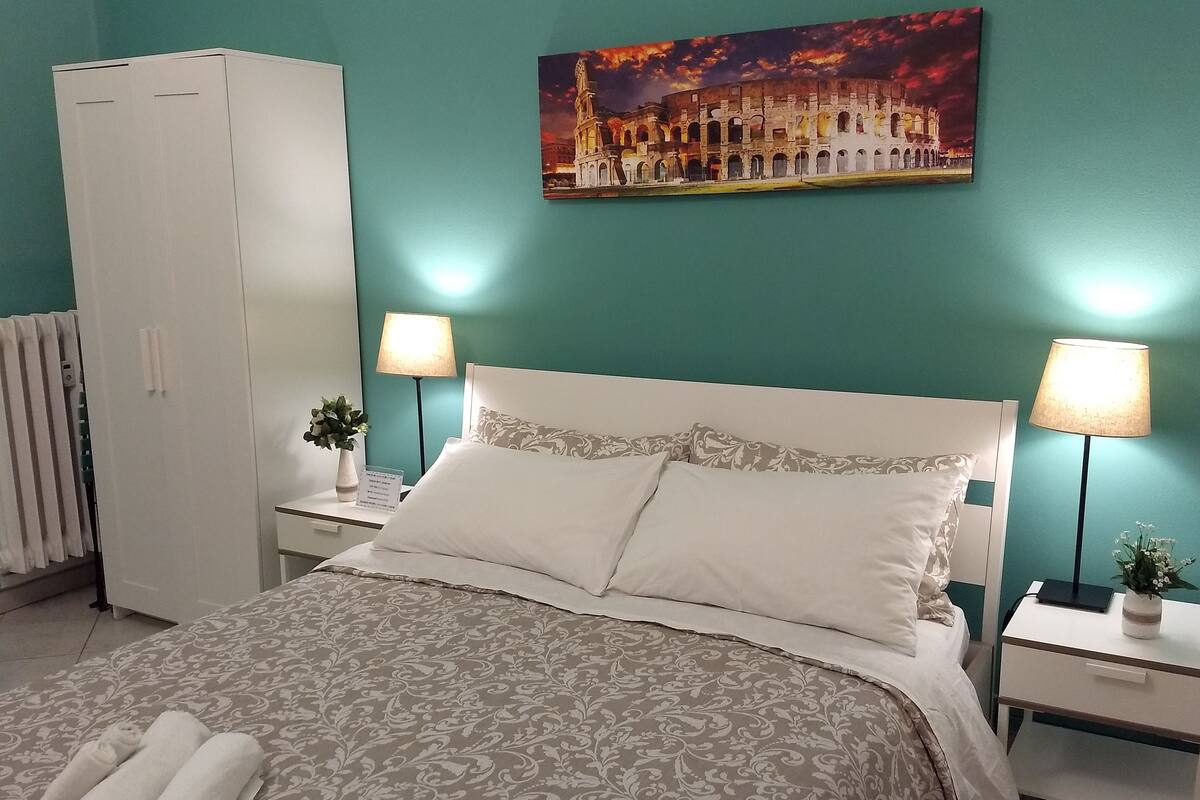A neatly made bed with a patterned bedspread, two bedside lamps, a nightstand with a small plant, and a large painting of a historical building on the wall.