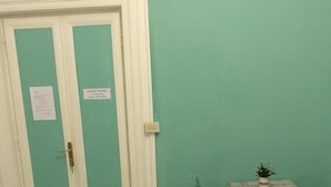 Quarto com paredes verde-água, uma porta branca com a placa 'Silence Hours', uma mesinha com uma planta e um sofá estampado.