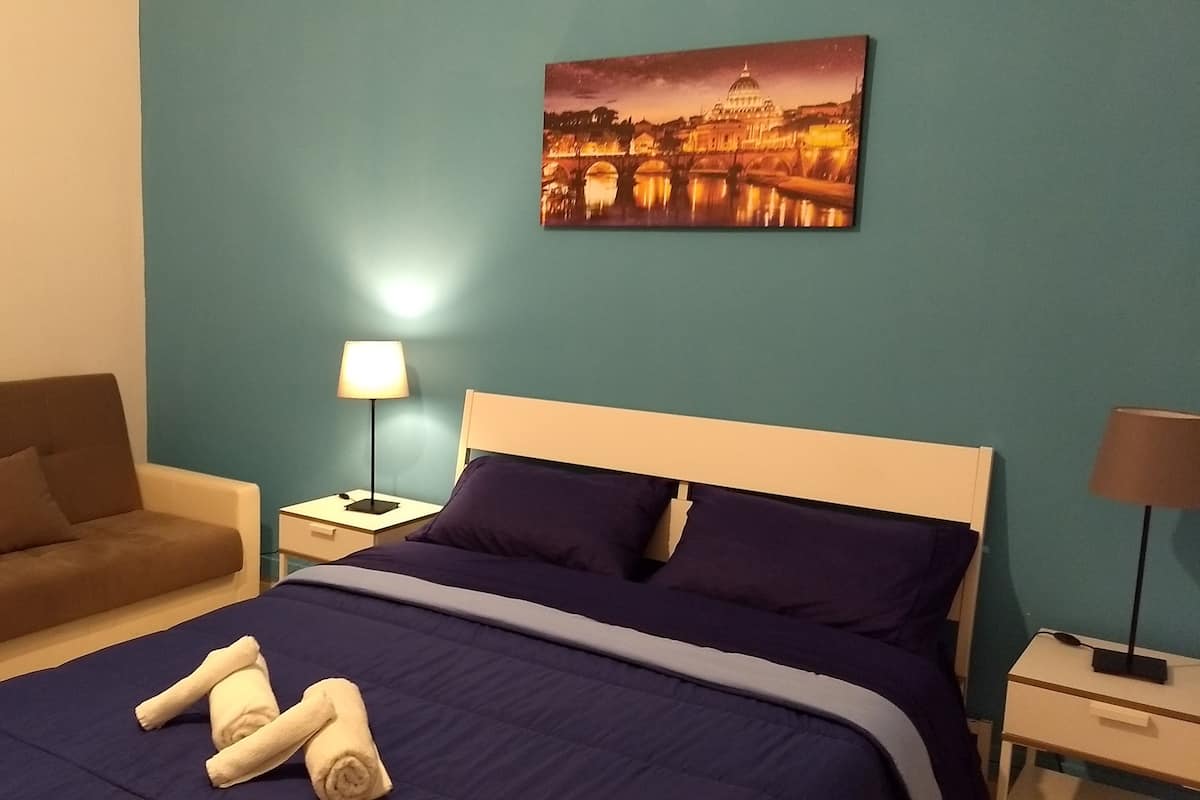 A hotel room with a bed, a nightstand, a lamp, a sofa, and a painting of a cityscape.