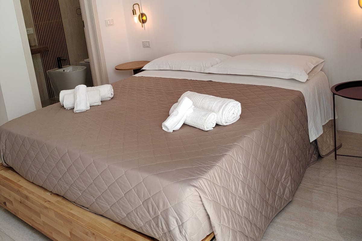 A modern bedroom with a large bed, a wooden floor, a bathroom with a bathtub, and two wall-mounted lights.