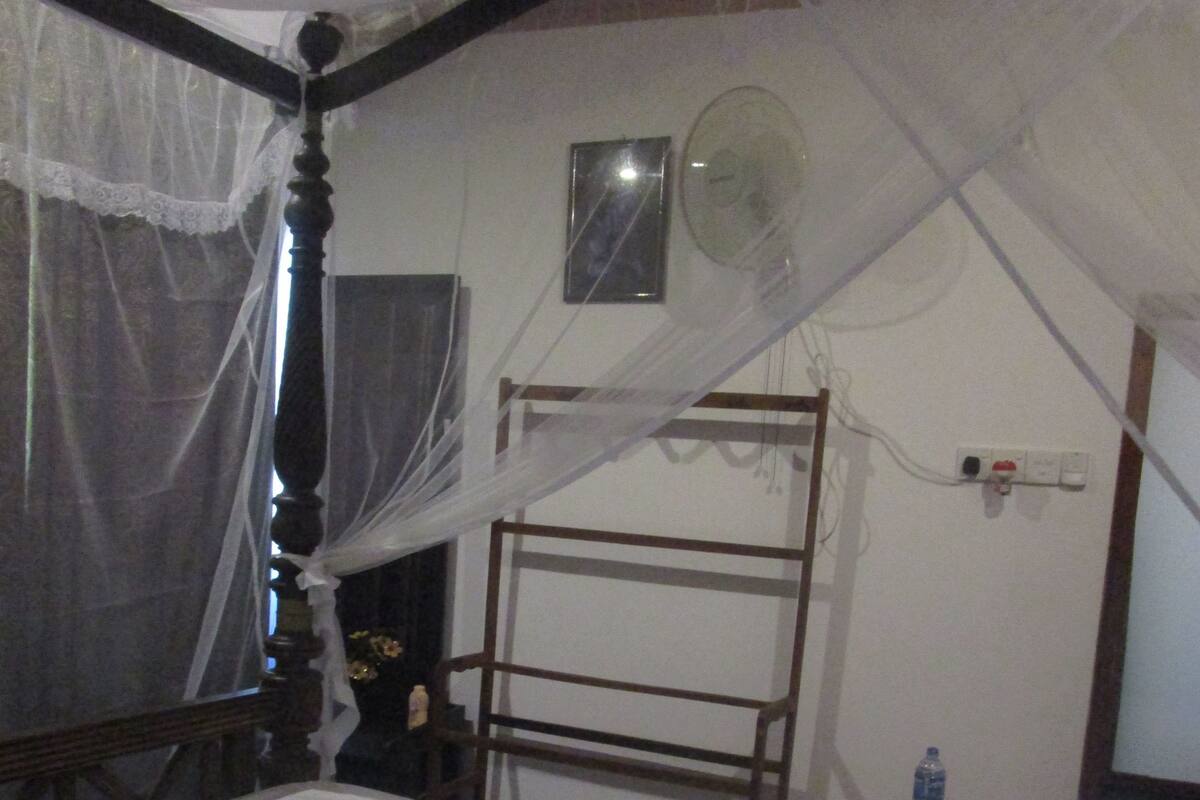 A room with a bed covered in white sheets, a bedside table with a lamp, and a wall socket.