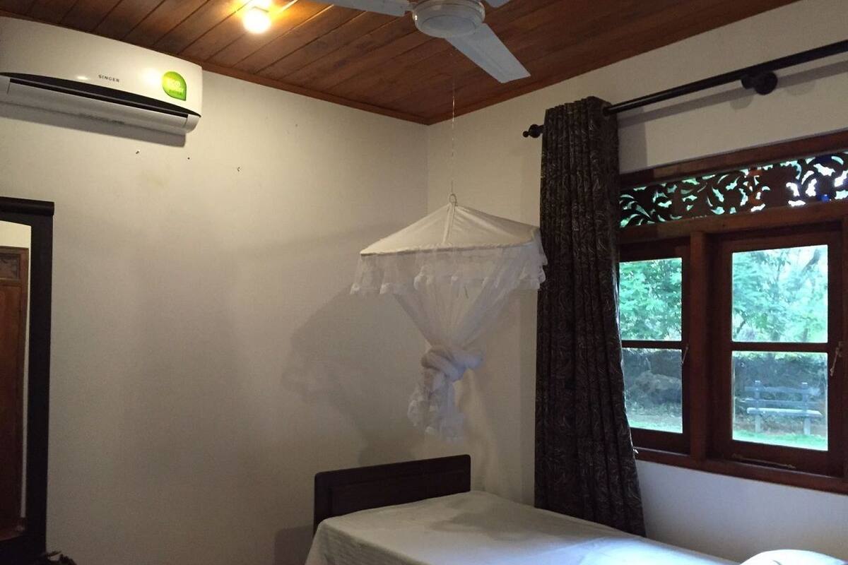 A small room with a single bed, a ceiling fan, an air conditioning unit, a window with a view of greenery, and a curtain.