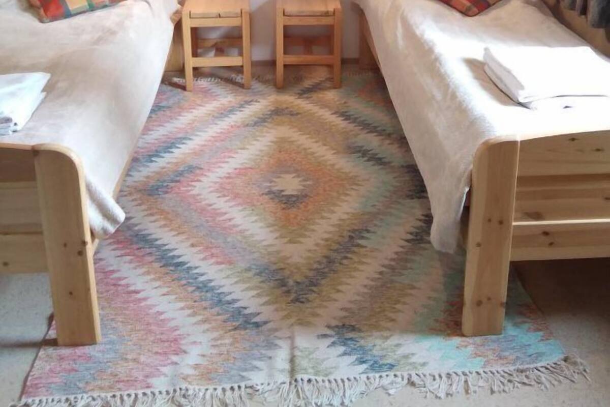 A small, cozy room with two wooden beds, a small wooden nightstand, a colorful rug, and a wall hanging above the beds.