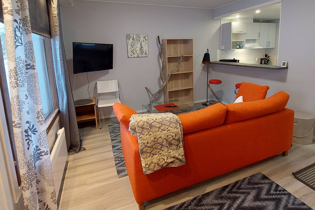 A modern living room with a bright orange sofa, a flat-screen TV, and a kitchen area with a large mirror.