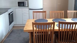 Een kleine keuken met witte kasten, een magnetron, een koelkast en een houten eettafel met stoelen.