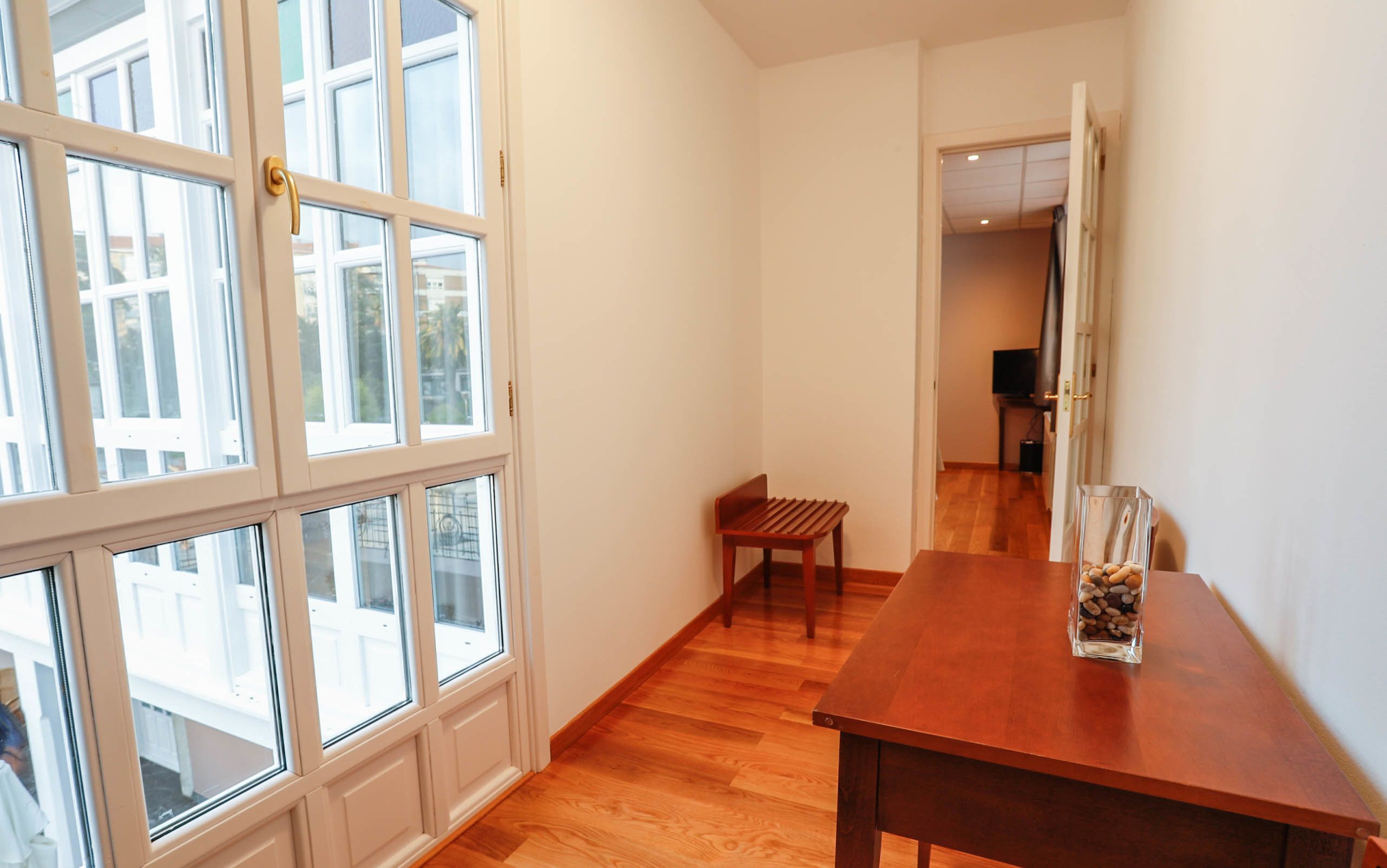 A narrow hallway with wooden flooring, a wooden bench, and a table with a vase of pebbles.